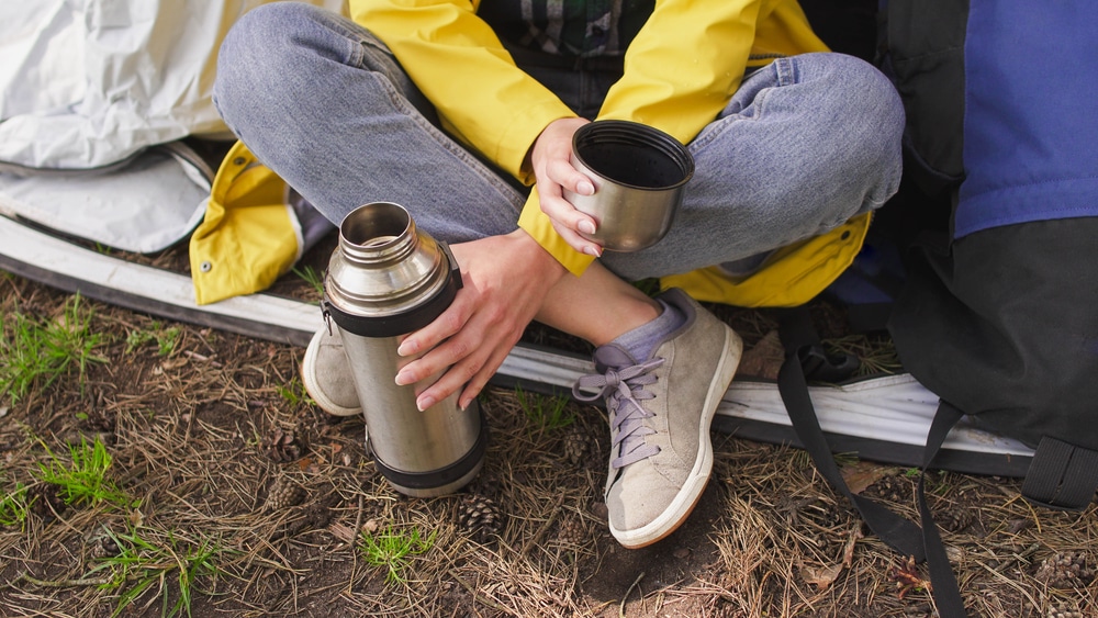 A woman sits near a tent and holding a thermos